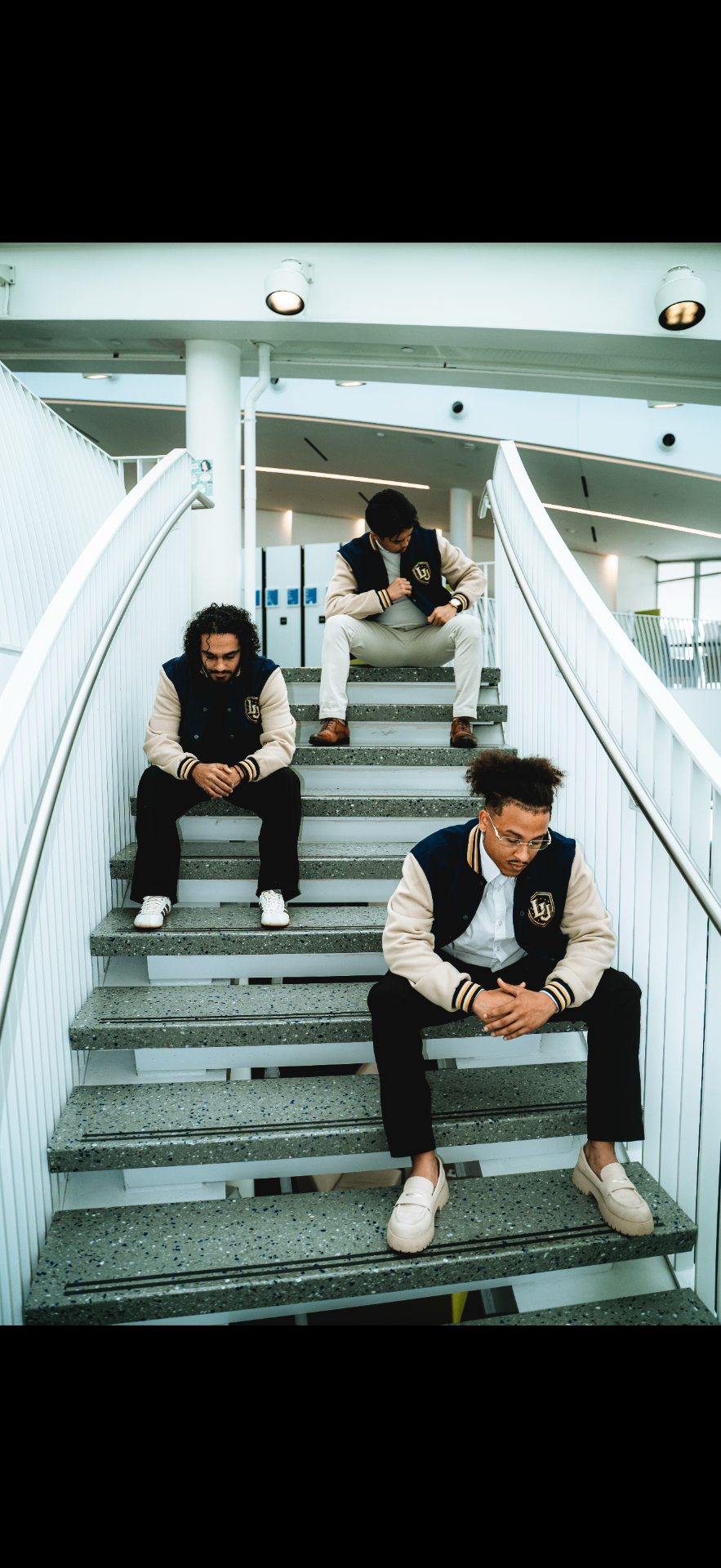 Three men wearing the Legends University varsity jacket, seated on an architectural staircase